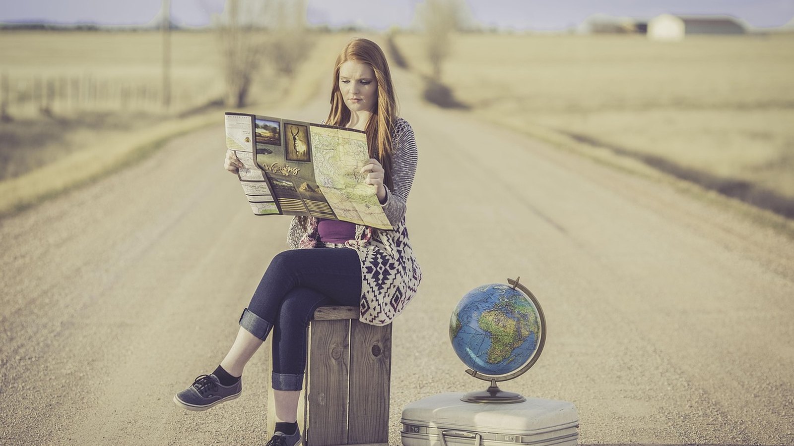 Žena s mapou sedí na starom kufri pri ceste, vedľa glóbus