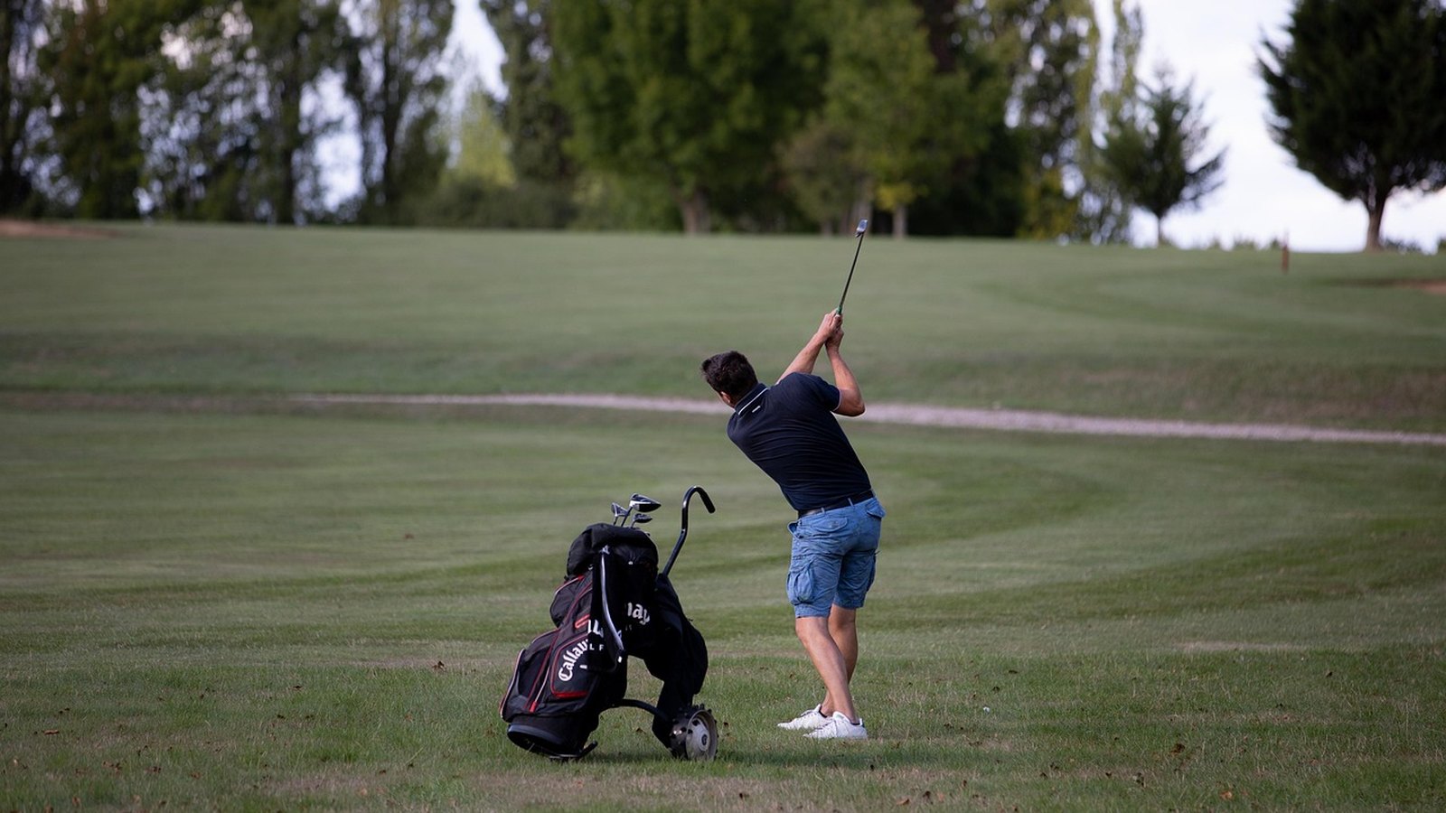 Golfista odpaľuje loptičku na ihrisku vedľa vozíka s palicami