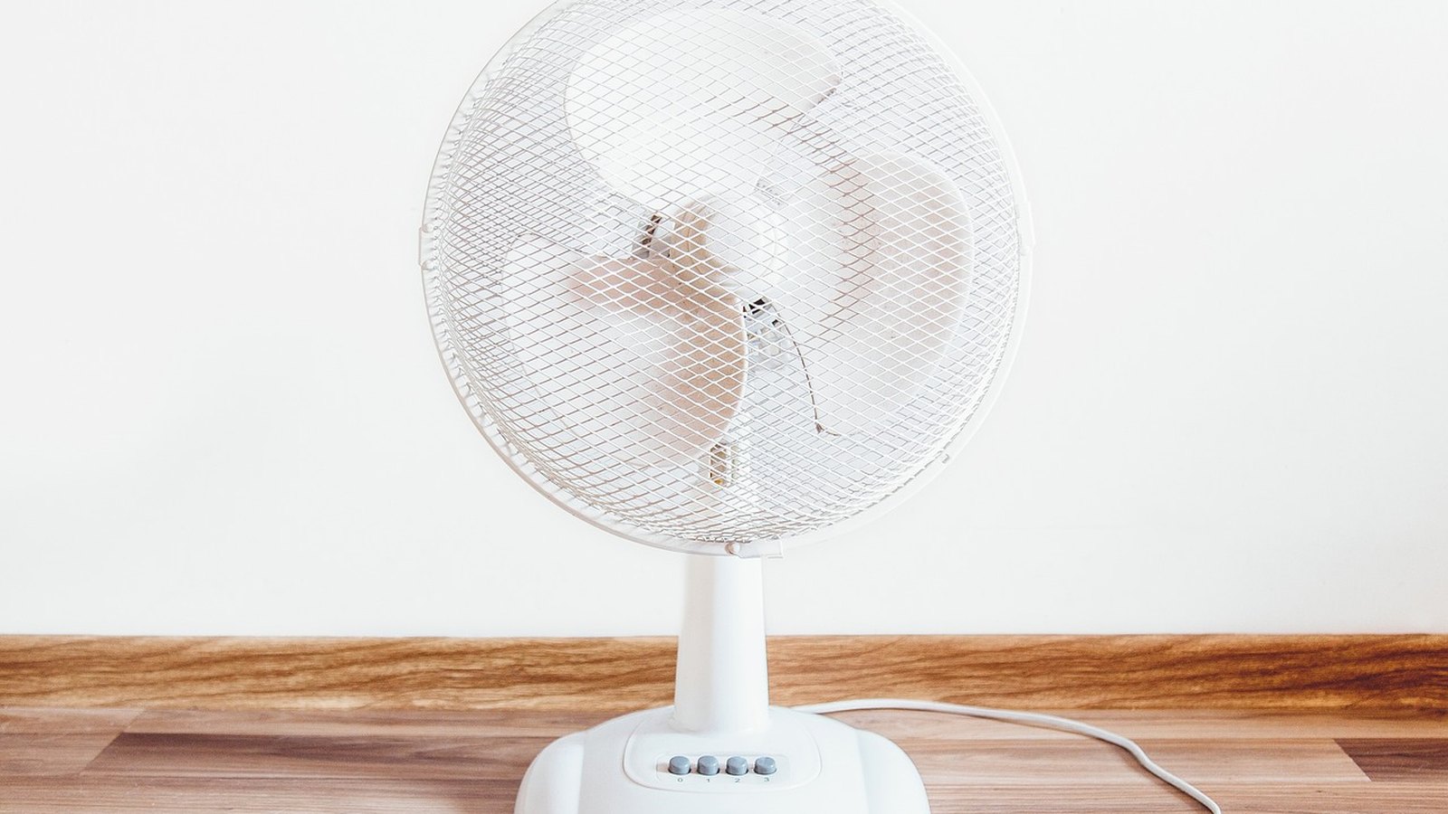 Biely stolový ventilátor na drevenej podlahe pri bielej stene