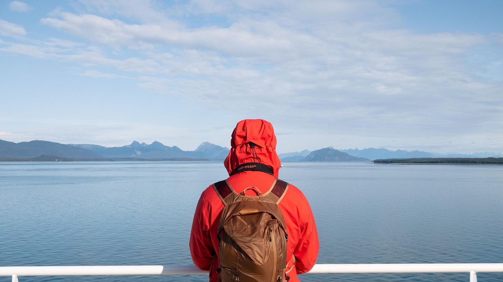 Turista v červenej bunde s hnedým batohom na pleciach pri mori